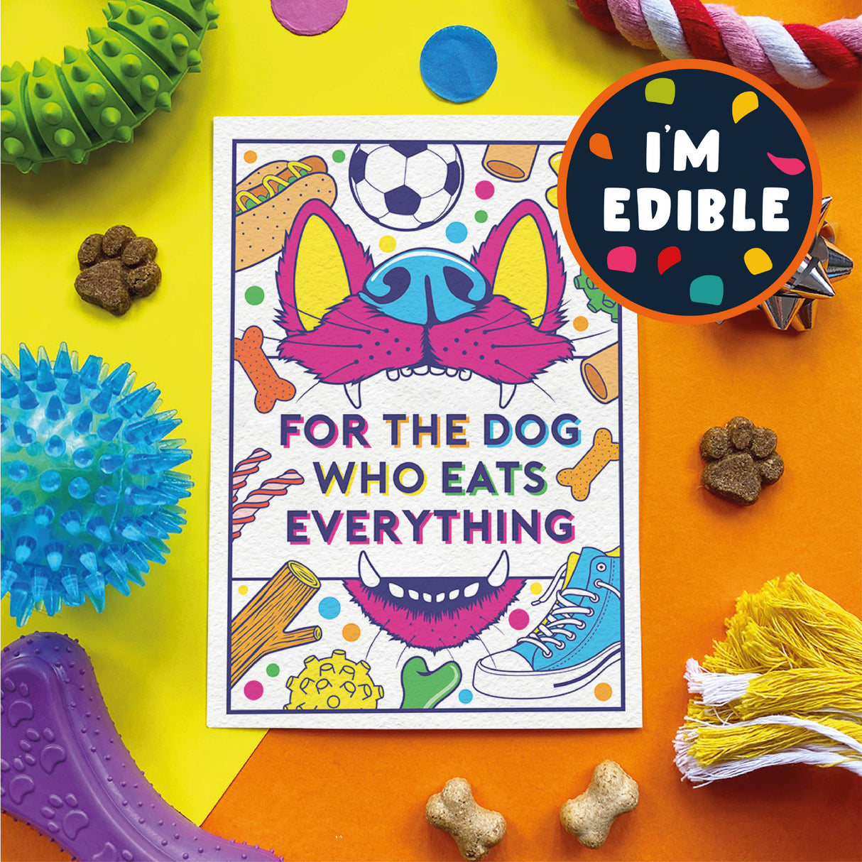 A square printed card or sticker with a colorful cartoon dog face and the text 'FOR THE DOG WHO EATS EVERYTHING,' surrounded by dog toys and treats on a bright yellow and orange background. A circular badge says 'I'M EDIBLE.