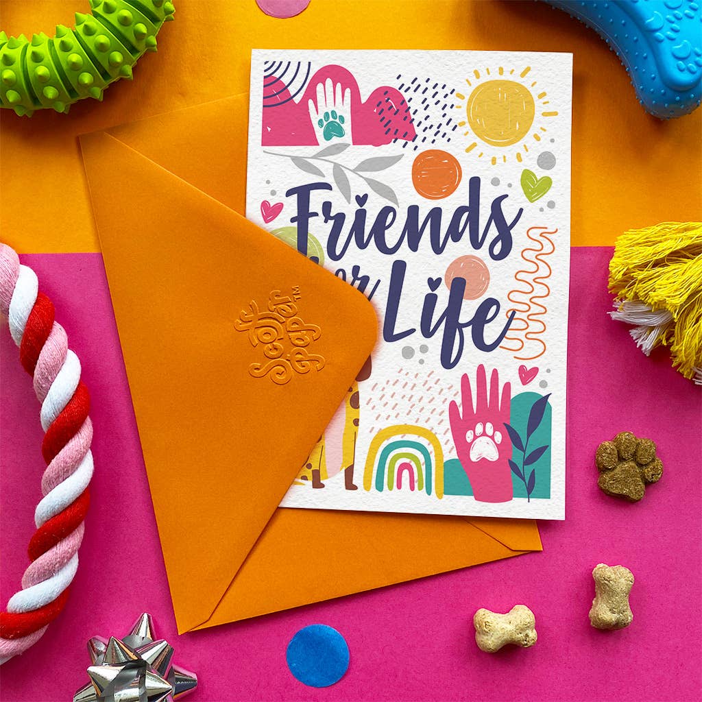 A colorful greeting card that reads 'Friends for Life' is displayed with an orange envelope on a bright, multi-colored flat-lay background with dog toys and treats.
