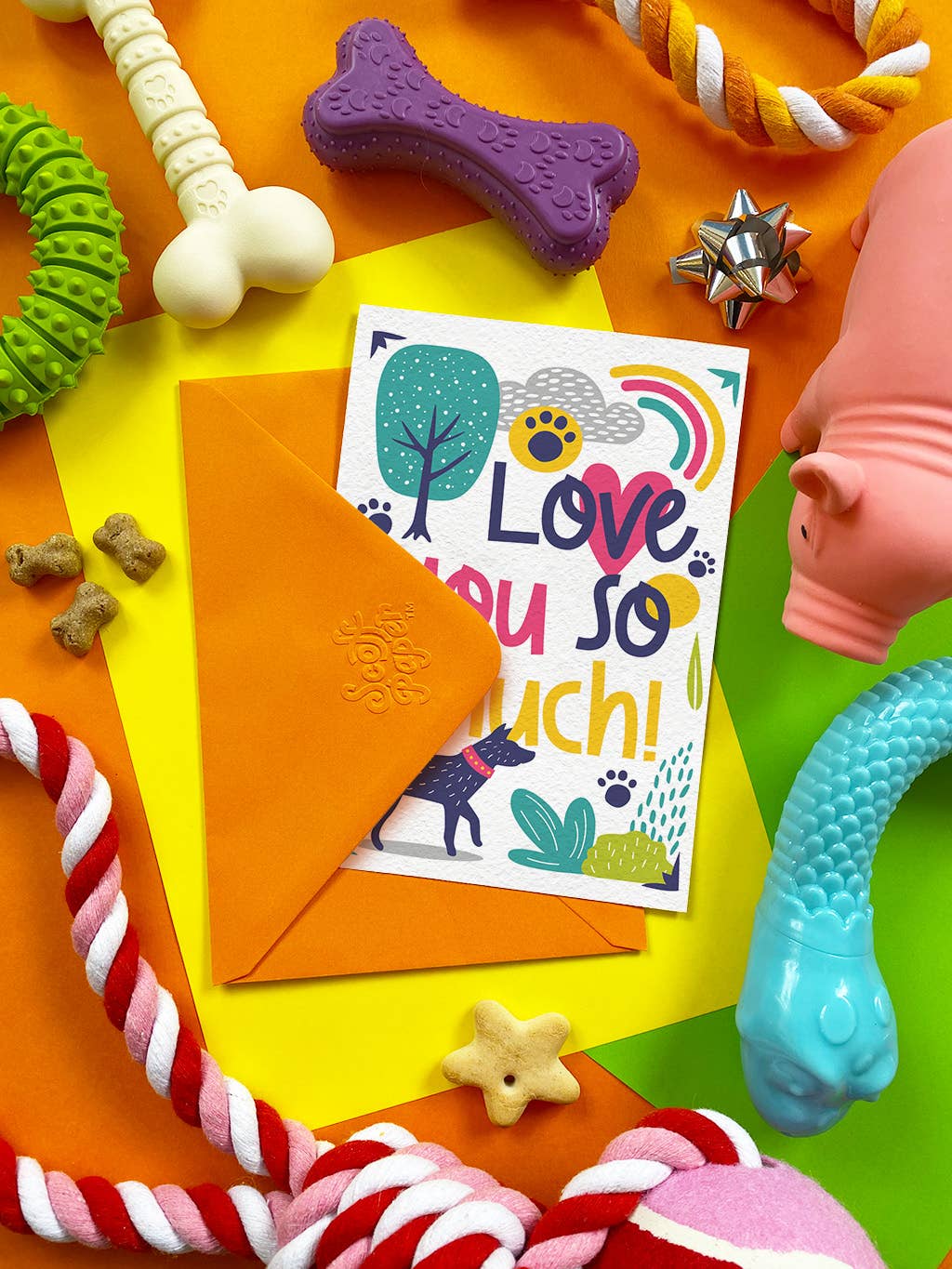 A square greeting card with colorful dog-themed illustrations and the text 'Love you so much!' is displayed on a bright yellow sheet, partially tucked into an orange envelope. The card is surrounded by colorful dog toys on a multicolored background.