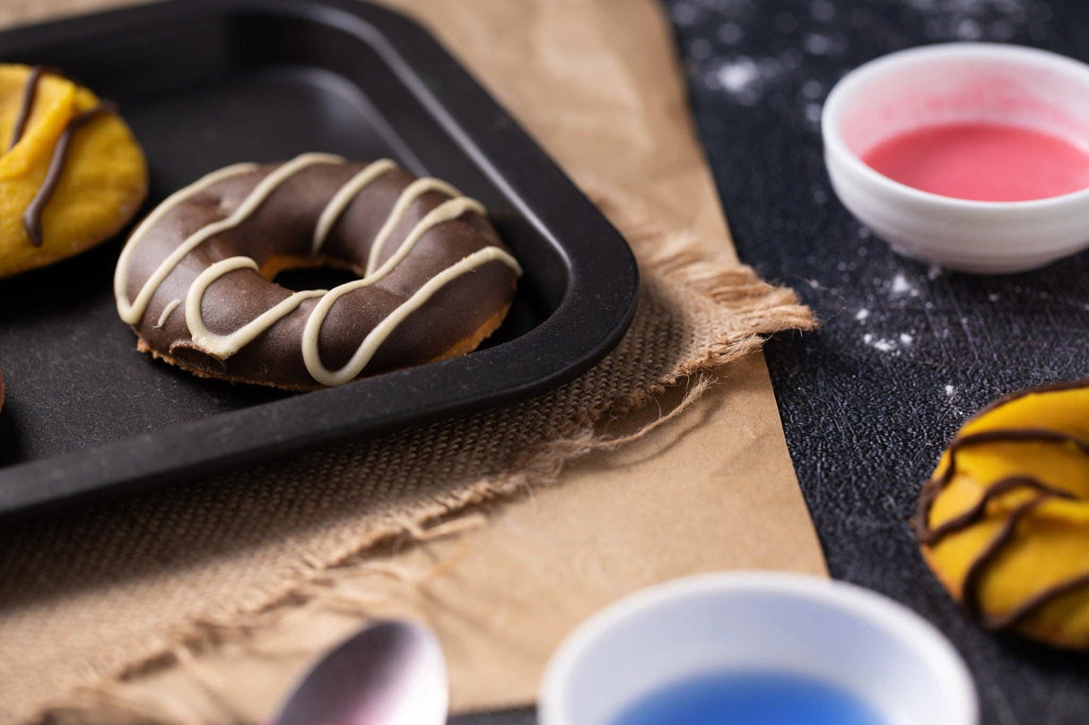 Chocolate dog donut with white drizzle on a black tray, surrounded by colorful donuts and small bowls of icing.
