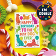 A colorful dog-themed birthday card reading 'HAPPY BIRTHDAY TO THE BEST DOG EVER' is shown on a bright yellow surface surrounded by dog toys and treats. A round badge in the top right says 'I'M EDIBLE'.