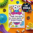 A square printed card or sticker with a colorful cartoon dog face and the text 'FOR THE DOG WHO EATS EVERYTHING,' surrounded by dog toys and treats on a bright yellow and orange background. A circular badge says 'I'M EDIBLE.