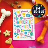A colorful printed card reading “FAVOURITE CHILD” surrounded by playful dog toys and treats on bright orange, pink, and red surfaces. A round badge says “I'M EDIBLE.