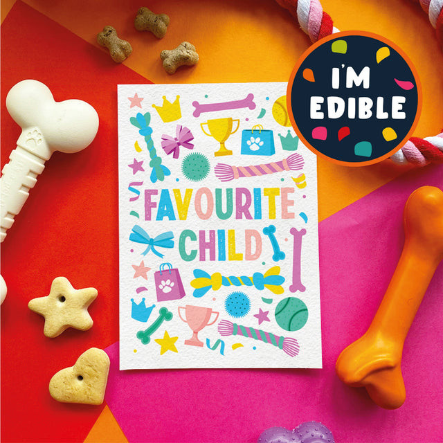 A colorful printed card reading “FAVOURITE CHILD” surrounded by playful dog toys and treats on bright orange, pink, and red surfaces. A round badge says “I'M EDIBLE.