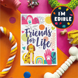 A square printed card or sticker with the phrase “Friends for Life” in large navy script, surrounded by colorful dog-themed illustrations including a yellow dog, pink paw prints, hearts, and a rainbow. The item is photographed in a styled flat lay on bright orange and pink backgrounds with rope toys, a bone-shaped toy, tassel, and a circular badge reading “I’m Edible.