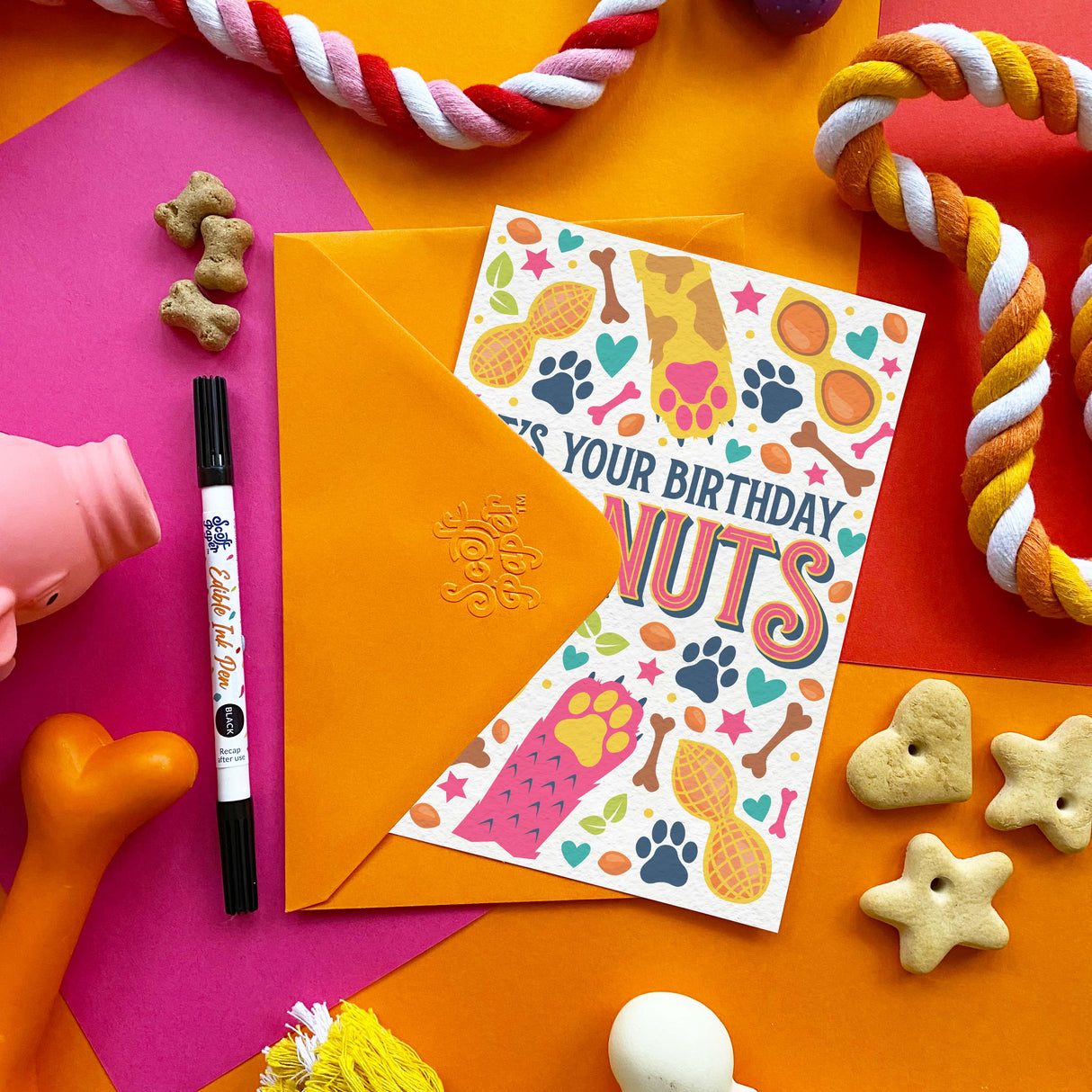A birthday greeting card with an orange envelope, styled in a colorful flat-lay with dog toys, treats, and a marker on a bright geometric background.