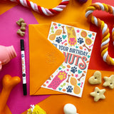 A birthday greeting card with an orange envelope, styled in a colorful flat-lay with dog toys, treats, and a marker on a bright geometric background.