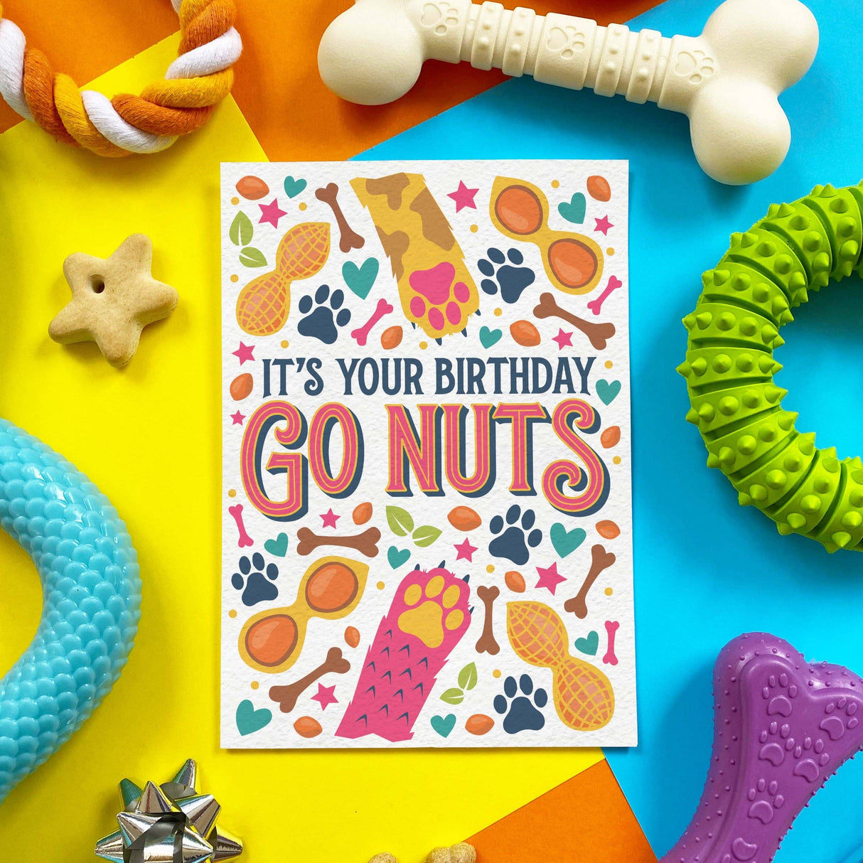 A square greeting card with a colorful dog-themed design reading 'IT'S YOUR BIRTHDAY GO NUTS,' surrounded by dog toys on a bright yellow, blue, and orange background.