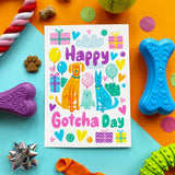 A colorful 'Happy Gotcha Day' dog and cat illustration print centered on a bright blue background, surrounded by playful dog toys including a blue bone-shaped toy, a purple chew, a green squeaky ball, and other party-themed accessories.