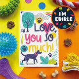 A square white card with colorful dog-themed artwork reading 'Love you so much!' is shown on a bright yellow and orange background with pet toys and treats around it. A round badge in the top right says 'I'M EDIBLE'.